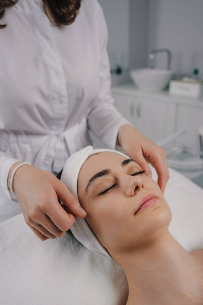 Cosecha femenina anónima aplicando mascarilla facial en la cara de una clienta joven con los ojos cerrados durante la rutina de cuidado de la piel en la mañana en casa - Foto, Imagen