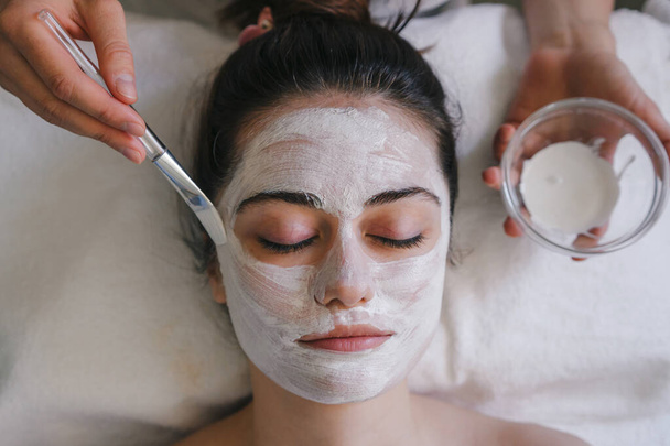 Draufsicht der Ernte anonyme weibliche Anwendung Gesichtsmaske auf Gesicht mit Pinsel, während im hellen Raum sitzen - Foto, Bild