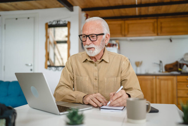 Ένας ηλικιωμένος καυκάσιος παππούς με γενειάδα και γυαλιά εργάζονται σε φορητό υπολογιστή χρήση στο σπίτι σε απευθείας σύνδεση ευτυχής χαμόγελο επιτυχία - Φωτογραφία, εικόνα