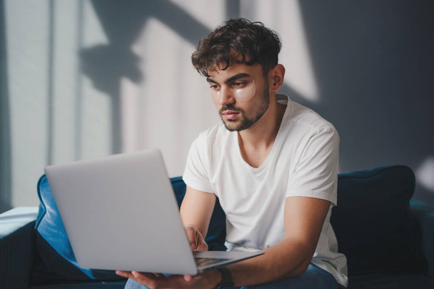 Concentrated male freelancer in casual clothes sitting on sofa and browsing laptop while working on remote project at home - 写真・画像