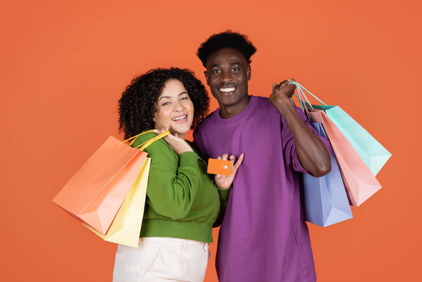 Šťastný veselý mladý multietnický pár s barevnými nákupními taškami a bankovní karty izolované na oranžové pozadí studia, černošky a hispánský dáma doporučující bezkontaktní nakupování, kopírovací prostor - Fotografie, Obrázek