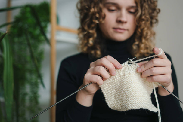 Crop young ethnic female with curly hair in casual clothes knitting with yarn and needles while working in light room at home - Foto, Bild