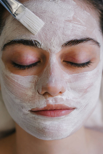 Crop jonge vrouw met gesloten ogen aanbrengen van gezichtsmasker op gezicht met borstel - Foto, afbeelding