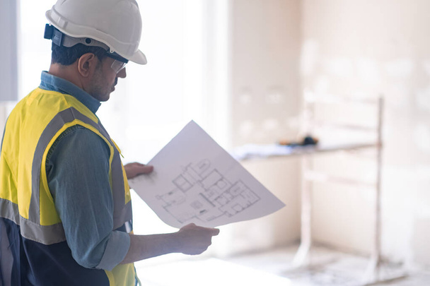Focused architect with apartment drawing in hands and construction safety equipment man in protective helmet and vest reads details standing in light empty room - Fotó, kép