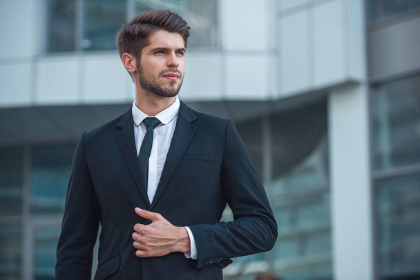 Handsome young confident businessman in classic suit is looking away, standing outside the office building - Fotoğraf, Görsel