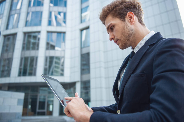 Side view of attractive young confident businessman in classic suit using a tablet, standing outside the office building - Foto, Bild