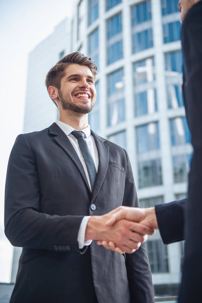 彼の同僚と握手と笑顔、オフィスビルの外に立って古典的なスーツでハンサムな青年実業家のローアングル ビュー - 写真・画像