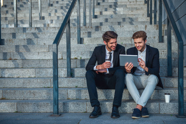 Two handsome young businessmen in formal clothes are using a tablet and smiling while sitting on stairs of the office building during coffee break - Foto, imagen
