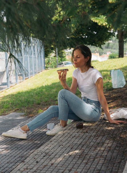 Woman sitting on the sidewalk spreading cocoa cream on a slice of bread for snack - Photo, Image