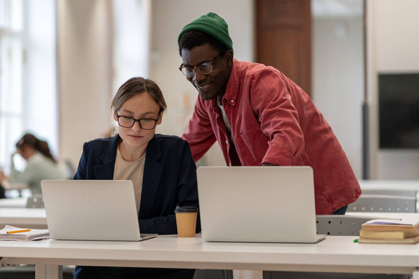 Een glimlachende Afrikaan helpt blanke meisjes die aan een project werken in de universiteitsbibliotheek. Positieve man vrouw kijk naar laptop scherm uit te leggen studeren samen op de campus. - Foto, afbeelding