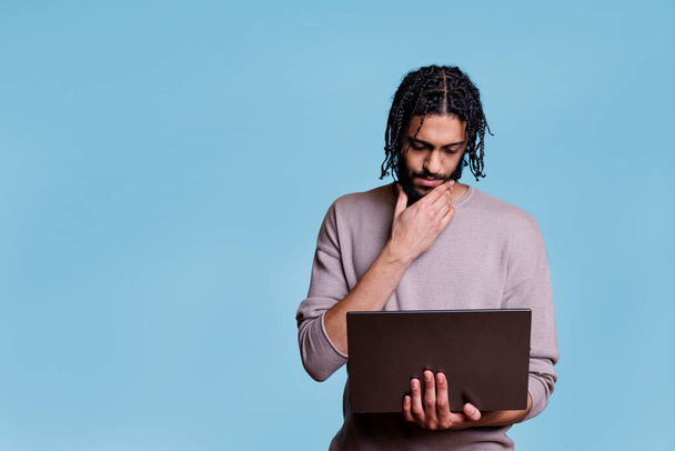 Młody, przemyślany arabski człowiek trzymający laptopa podczas analizy kodu oprogramowania i pocierania podbródka. Pensive osoba myśląca wątpliwość podczas czytania raportu biznesowego na komputerze przenośnym - Zdjęcie, obraz