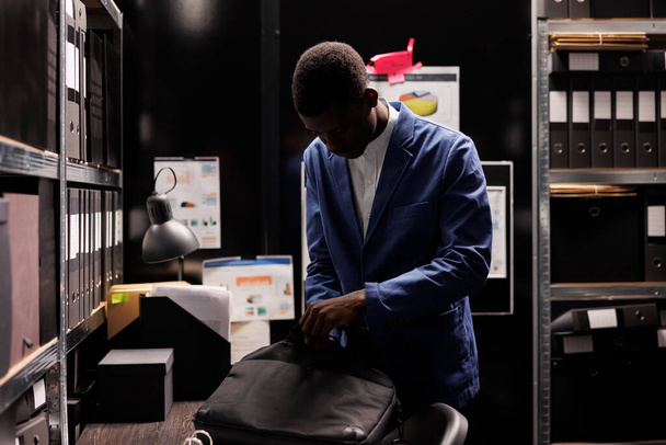 Afro-Amerikaanse boekhouder stopt spullen in zijn rugzak, verlaat het werk laat op de avond. Zakenman die overuren maakt in de administratie, administratieve documenten analyseert in de opslagruimte. - Foto, afbeelding
