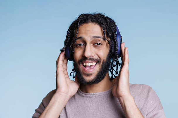 Glücklicher arabischer Mann, der fröhlichen Liedern in drahtlosen Kopfhörern lauscht. Lächelnder Aufgeregter genießt Gesang im Kopfhörer und blickt mit freudigem Gesichtsausdruck in die Kamera - Foto, Bild