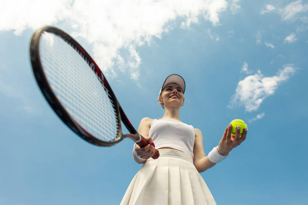 dívka tenista v bílé uniformě drží raketu a míč na tenisovém kurtu, žena sportovec hraje tenis venku proti obloze - Fotografie, Obrázek