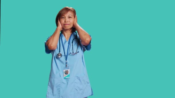 Sertifioitu sairaanhoitaja, jota vaivaa liiallinen melu töissä. Terveydenhuollon asiantuntija peittää korvat kädet, harmittaa äänekäs ympäröivä ääniä, eristetty sininen studio tausta - Materiaali, video
