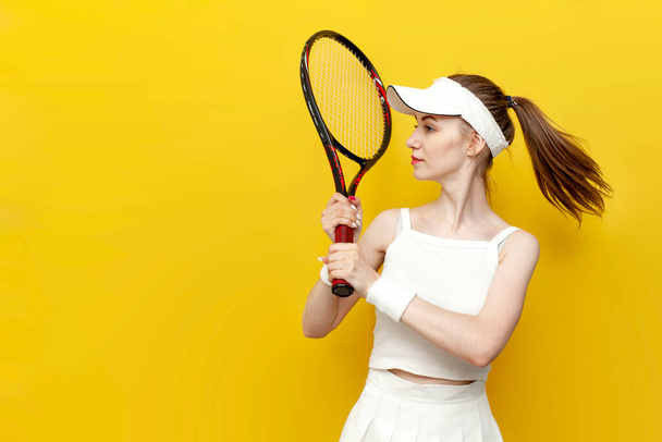 girl tennis player in sportswear holding tennis racket on yellow isolated background and smiling, portrait of female tennis coach in white uniform with sports equipment - Photo, Image