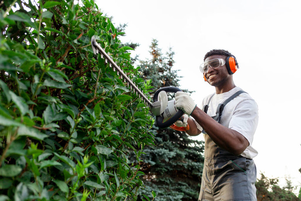 trabajador de jardín masculino afroamericano en uniformes recorta arbustos con herramienta eléctrica, hombre con gafas y auriculares trabaja con cortador de cepillos y recorta árboles y vegetación - Foto, imagen
