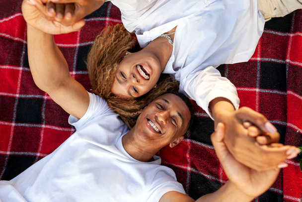 Африканская американская молодая пара лежит на одеяле и смотрит вверх и улыбается, мужчина с женщиной отдыхает и держит за руки, вид сверху - Фото, изображение