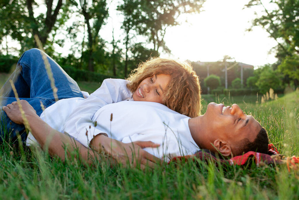 Αφροαμερικανοί ρομαντικό ζευγάρι βρίσκεται στο γρασίδι στο πάρκο και κοιμάται στη φύση, ευτυχισμένη οικογένεια ξεκουράζεται και όνειρα σε εξωτερικούς χώρους, γυναίκα αγκαλιάζει το φίλο της και χαμογελά - Φωτογραφία, εικόνα