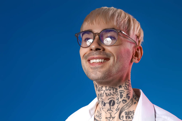 Mladý tetovaný muž v brýlích na modrém pozadí, detailní záběr - Fotografie, Obrázek