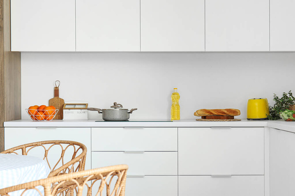 Λευκοί μετρητές με ηλεκτρική κουζίνα, κατσαρόλα, τοστιέρα και καλάθι με φρούτα στη σύγχρονη κουζίνα - Φωτογραφία, εικόνα
