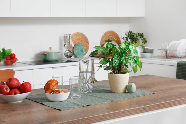 Houten eilandtafel met kamerplant, appels en kaneelbroodjes in moderne keuken - Foto, afbeelding