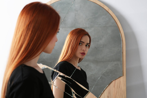 Mental problems. Depressed young woman looking at herself in broken mirror indoors - 写真・画像