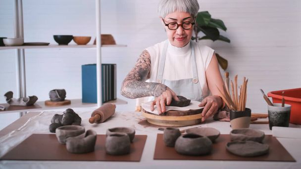自宅で陶芸作品を楽しむアジアの高齢女性。女性陶芸家が工房で新しい陶器を作っています. - 写真・画像