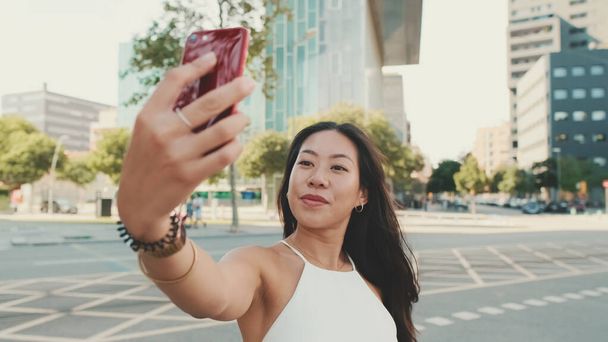 Cute happy girl dressed in white top and jeans makes selfie on mobile phone while standing on modern city background - Фото, изображение