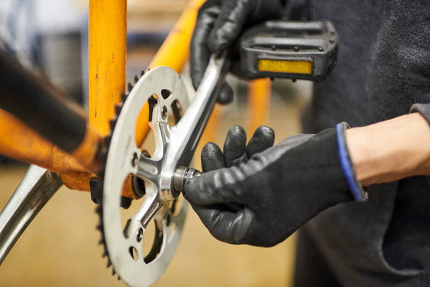 Уход за велосипедом: руки неузнаваемого человека в перчатках разбирают педали оранжевого велосипеда в мастерской. - Фото, изображение