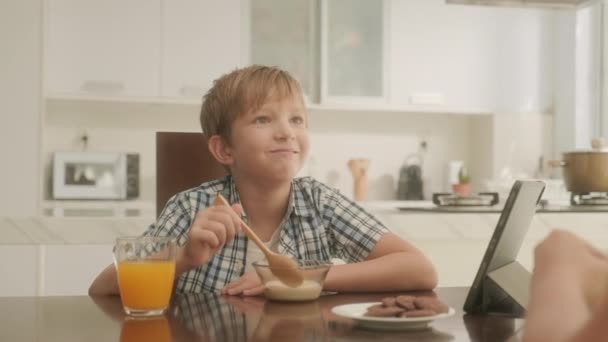 Маленький сын наслаждается завтраком с кукурузными хлопьями, соком и печеньем, разговаривая с матерью и смотря мультфильм на цифровом планшете - Кадры, видео