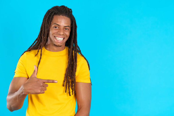 Happy latin man with dreadlocks pointing a blank space to the side in studio with blue background - Fotoğraf, Görsel