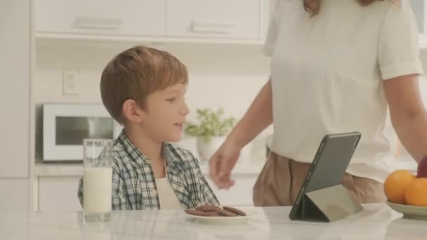 chłopiec oglądanie wideo na cyfrowy tablet siedzi w kuchnia stół z szklanką mleko mama całuje syn w policzek - Materiał filmowy, wideo