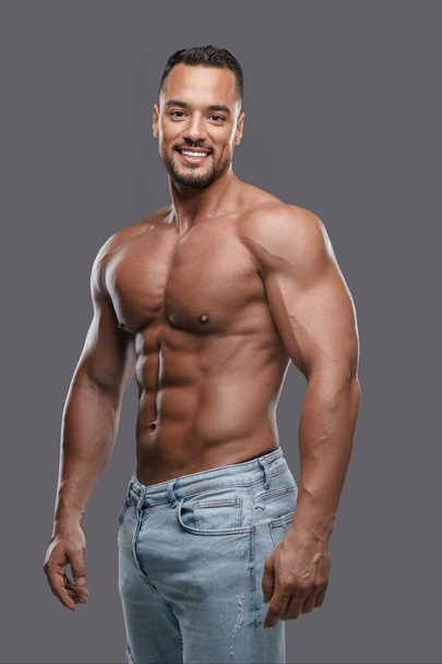 Charming and muscular male model with a dazzling smile poses shirtless against a neutral grey backdrop, emanating confidence and sex appeal - Фото, изображение