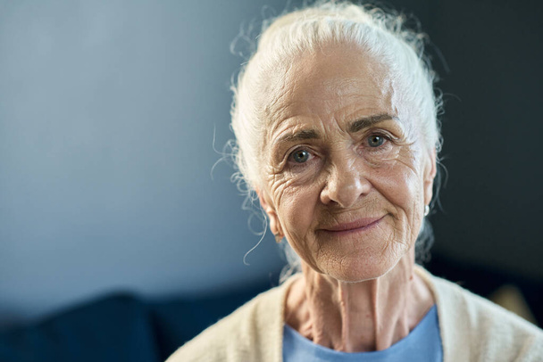 Twarz uśmiechniętej starszej kobiety z siwymi włosami patrzącej na ciebie z uśmiechem, siedzącej przed kamerą na niebieskim tle w izolacji - Zdjęcie, obraz