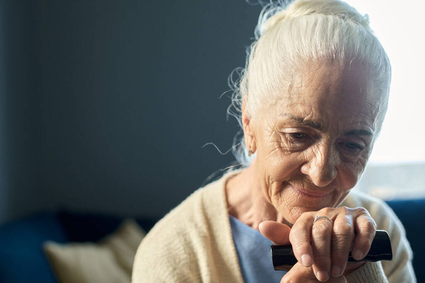 Donna anziana pensierosa con i capelli grigi che tiene la mano sulla maniglia del bastone da passeggio mentre si siede davanti alla telecamera e pensa a qualcosa di buono - Foto, immagini