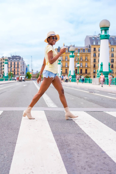 Portrait of black ethnic woman drinking an orange juice enjoying summer on vacation crossing the street at the zebra crossing - Photo, Image