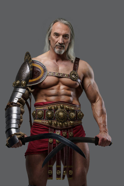 Distinguido gladiador anciano exuda potencia y fuerza en armadura elegante y ligera, empuñando dos espadas de gladius con confianza sobre un fondo gris - Foto, Imagen
