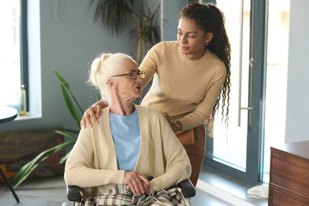 Hezké multietnické dospívající dívka stojící vedle své babičky na invalidním vozíku a poslouchat ji, zatímco oba tráví víkend spolu - Fotografie, Obrázek