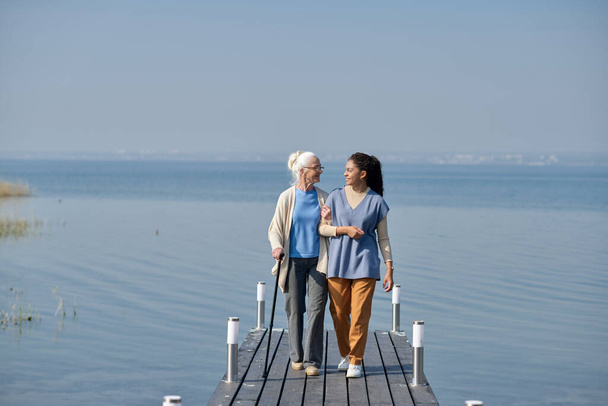 Lange kans van oude vrouw en haar kleindochter lopen langs pier tegen blauw meer en lucht en kijken elkaar aan terwijl ze kletsen - Foto, afbeelding