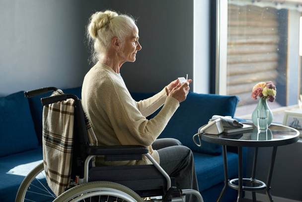 Oldalsó nézet a nyugdíjas nő fogyatékossággal fog venni tablettát kis műanyag tabletta-doboz, miközben ül tolószékben otthoni környezetben - Fotó, kép