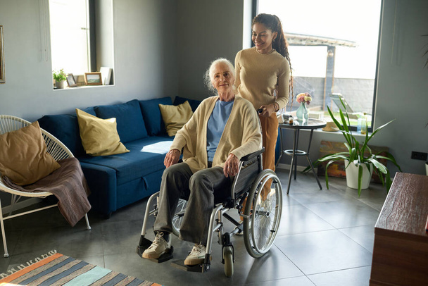 Teenage girl pushing wheelchair with her grandmother while walking along spacious living room with comfortable couch and armchair - Photo, Image