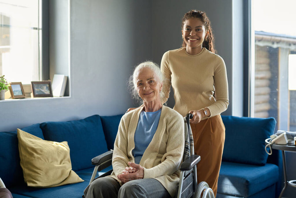 Cheerful multiethnic girl standing by her grandmather in wheelchair while both looking at camera with smiles against home environment - Foto, afbeelding