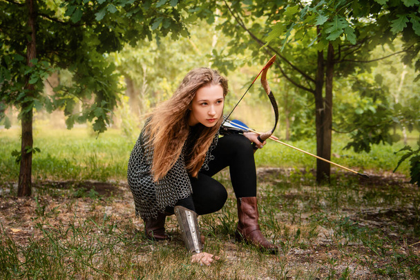 Medieval warrior woman in chain mail and bracers with a bow in her hands against the backdrop of an evening summer forest. - Фото, зображення