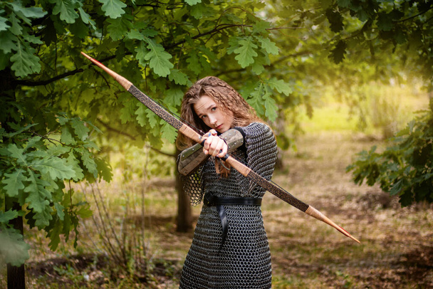 Medieval warrior woman in chain mail and bracers with a bow in her hands against the backdrop of an evening summer forest. - Фото, изображение
