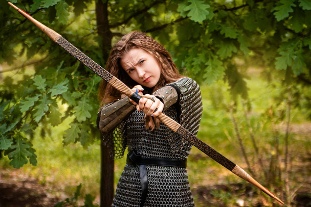 Medieval warrior woman in chain mail and bracers with a bow in her hands against the backdrop of an evening summer forest. - Fotografie, Obrázek