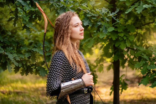 Medieval warrior woman in chain mail and bracers with a bow in her hands against the backdrop of an evening summer forest. - Fotoğraf, Görsel