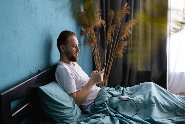 Young man lies in bed under blanket woke up after healthy sleeping holds smartphone check new messages in morning charged his mobile phone with power bank. Copy space - Foto, Bild