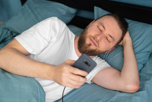 Atraktivní muž používající nabíjení chytrého telefonu na posteli, lidé a technologický koncept - Fotografie, Obrázek