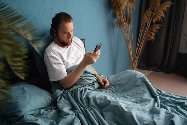 ヘッドフォンでベッドの上に寝そべって髭の男は、スマートフォンで好きな音楽を聞きます。一人で家でリラックスして - 写真・画像